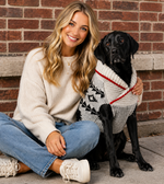 Woman sitting with a black dog wearing a sweater against a brick wall.