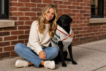 Woman sitting on the ground with a black dog wearing a sweater against a brick wall.